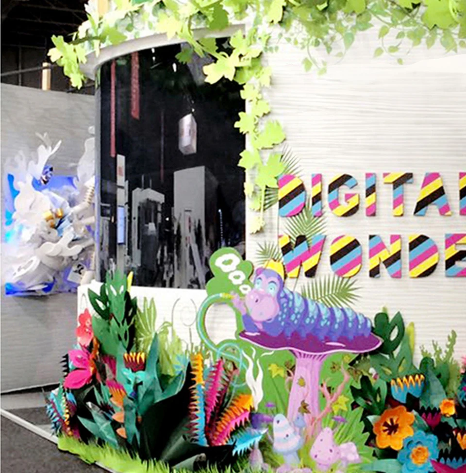 An exhibition stand lettered Digital Wonderland: a paper caterpillar on a mushroom among bright paper plants, the white Alice piece on the wall behind